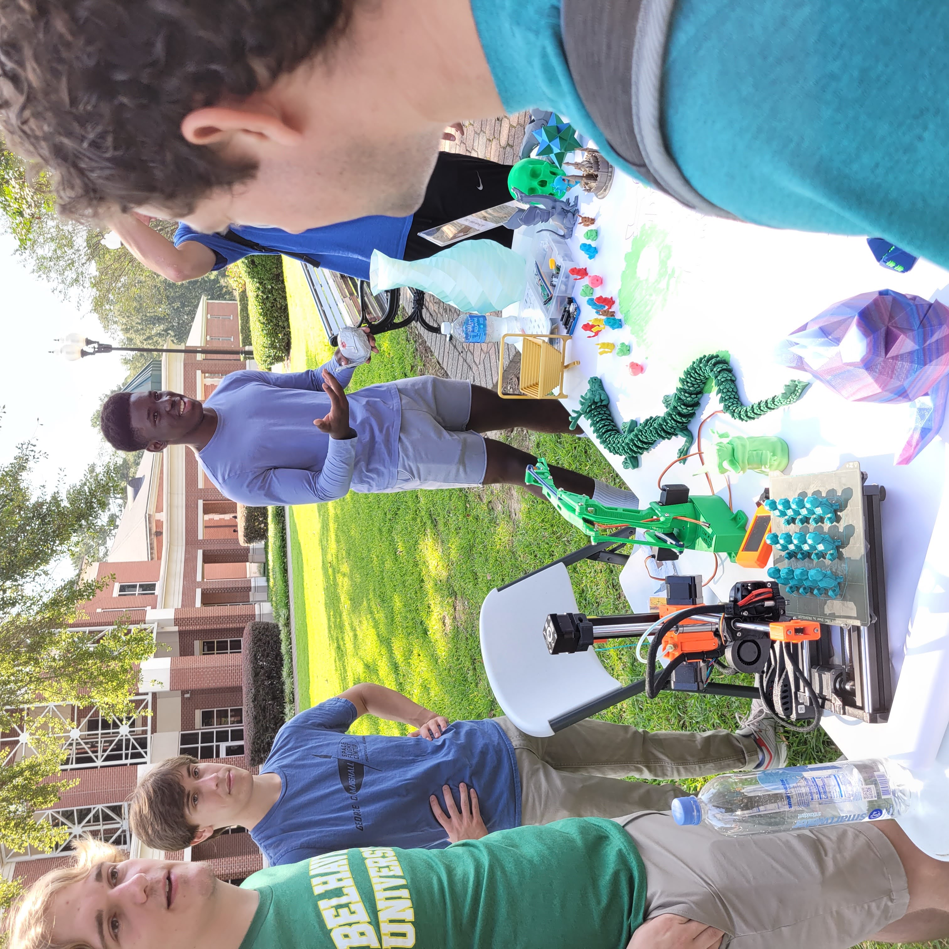 Four students stand around a folding table on a campus lawn. The table is covered with various colorful gadgets.