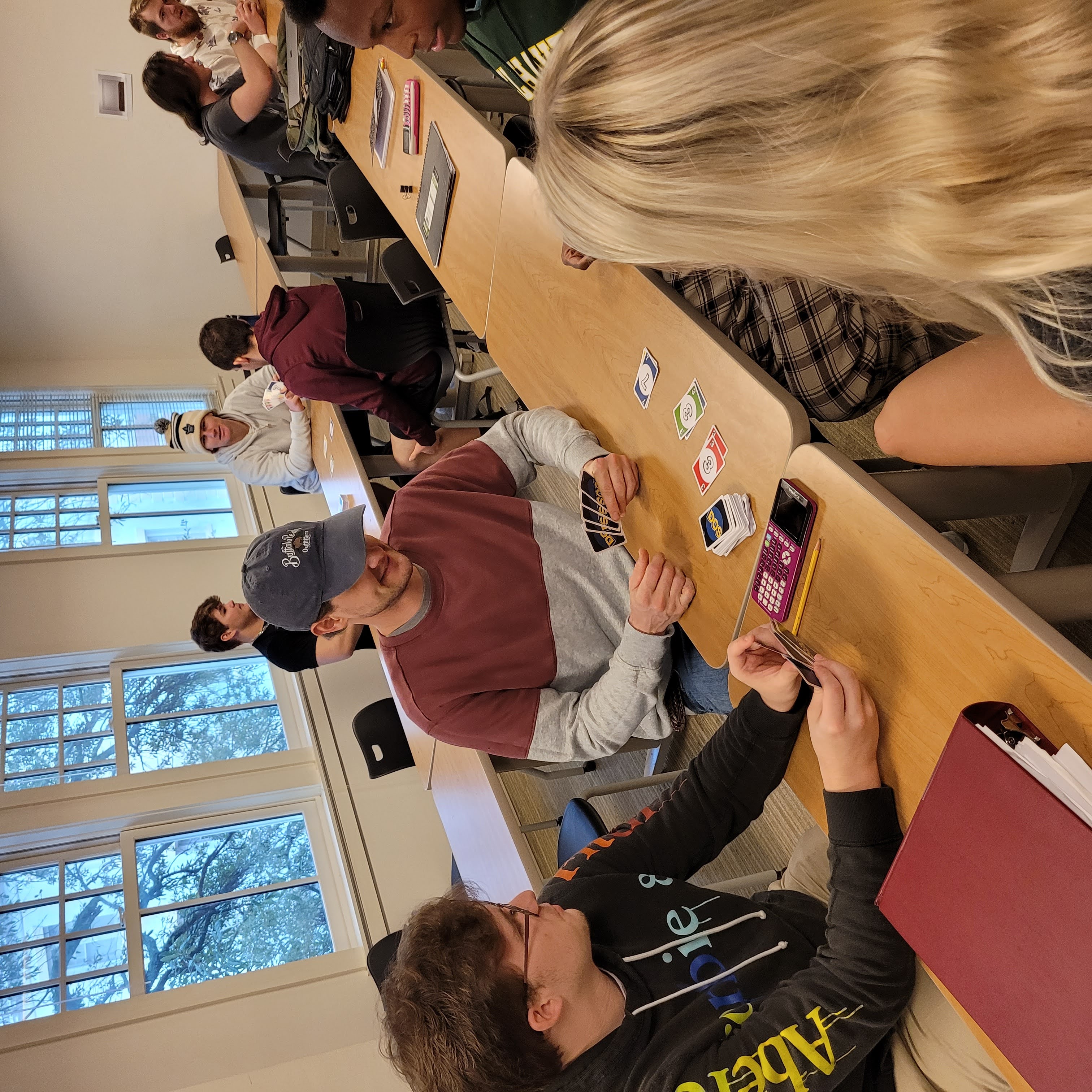 Several students sit in a classroom with long tables. Upfront are three young men and a woman holding cards that say "Dos" on their backs, similar to Uno playing cards.
