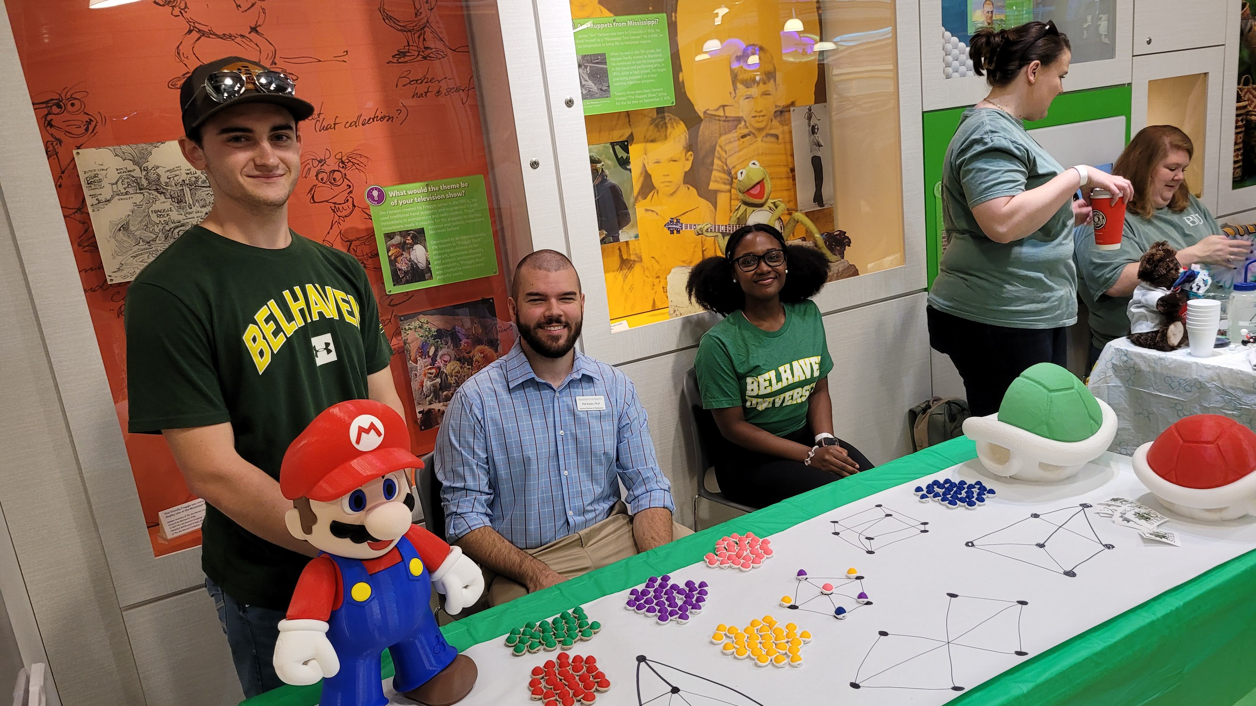 Several adults at a table with parchment paper. Several combinatorial graphs are drawn on the paper, with tokens positioned at some vertices. Plush Super Mario and two Koopa Shell dolls are at the table as well.