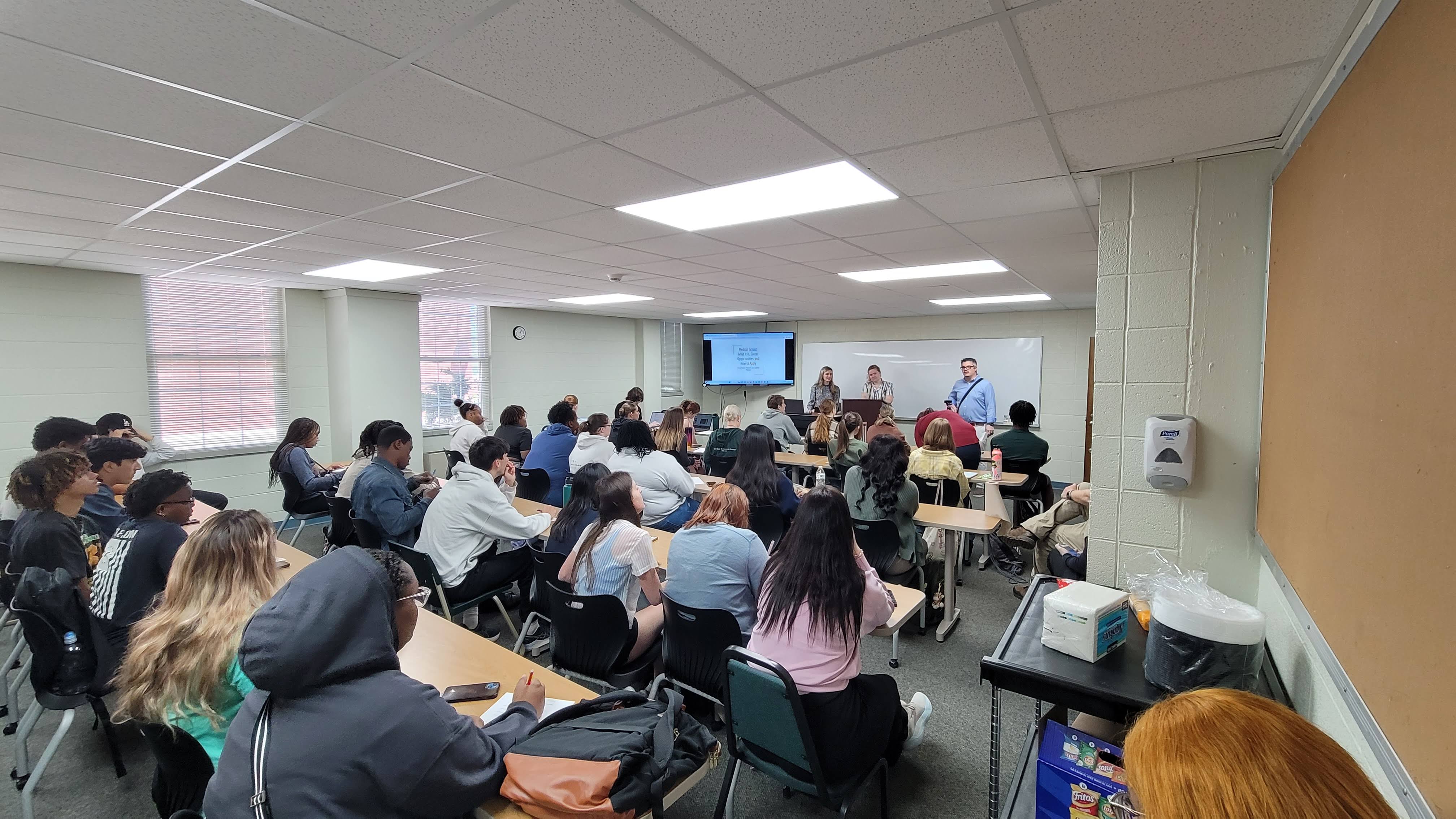 A classroom seated to capacity. A speaker stands at the front with a presentation displayed on a monitor.