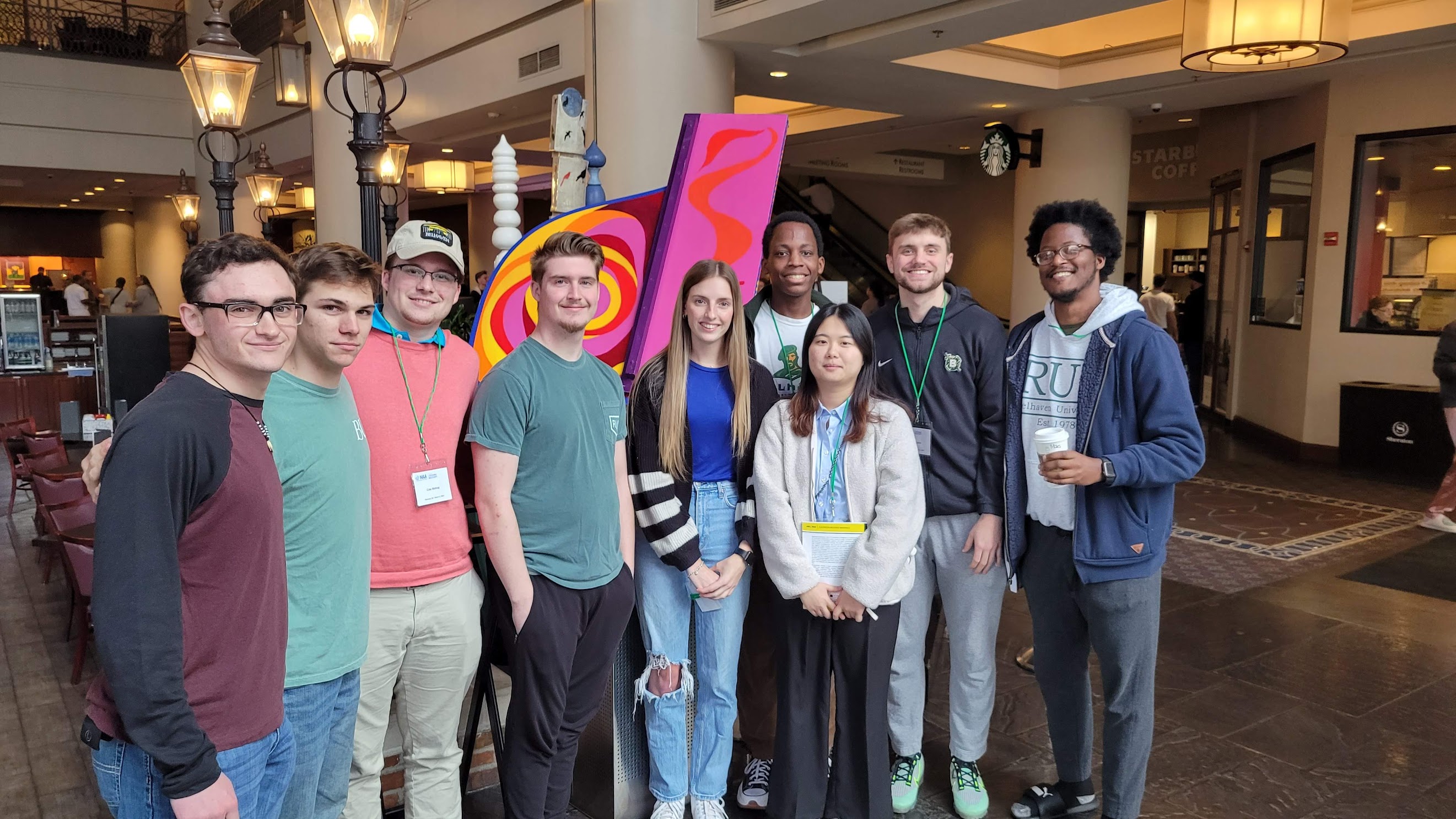 Nine college students of multiple races and genders stand together in an academic building hallways wearing nametags.